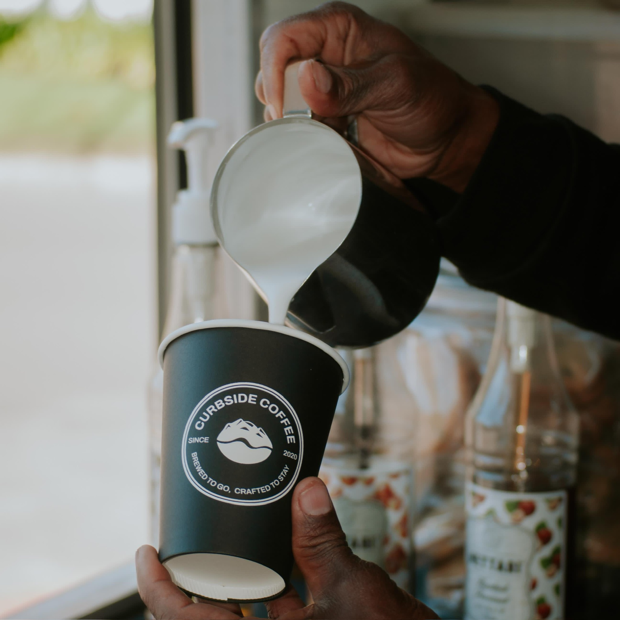 Curbside Coffee barista pouring milk into a branded cup at the service window